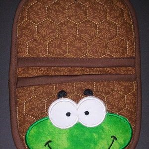 May include: Brown quilted pot holder with a frog face design. The pot holder features a brown background with a leaf pattern, a green frog face, and white eyes. It has a loop for hanging.