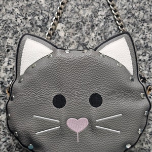 May include: A gray faux leather cat-shaped crossbody bag with white ears and a pink heart nose. The bag has a silver chain strap and is decorated with silver studs.