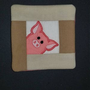 May include: Square fabric coaster featuring a pink pig design. The pig is embroidered on a white square, framed by tan and brown fabric. The coaster has a beige border and a pink edge.