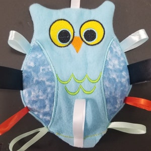 May include: A blue owl-shaped baby toy with embroidered eyes and a green belly. The toy has multiple ribbons attached to it in various colours, including black, white, orange, and light blue.