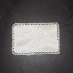 Puede incluir: Un parche rectangular blanco con un borde cosido gris.