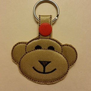 Może przedstawiać: Brązowy brelok z tkaniny w kształcie małpy z czerwonym guzikiem w kształcie serca i srebrnym kółkiem na klucze.