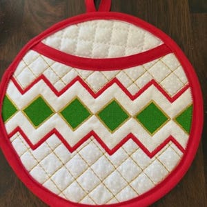 Może przedstawiać: Ochraniacz na garnki w kształcie czerwonej, białej i zielonej bombki choinkowej z wzorem diamentowym. Ochraniacz na garnki ma pętlę do zawieszenia.