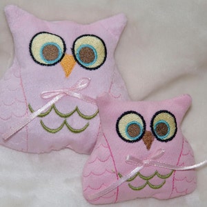 May include: Two pink plush owl toys with embroidered details. The larger owl has light blue eyes, a brown beak, and a pink bow. The smaller owl has light blue eyes, a brown beak, and a pink bow.