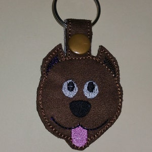 May include: Brown felt dog keychain with a pink tongue sticking out, black nose, and white eyes. The keychain has a gold button and a silver key ring.