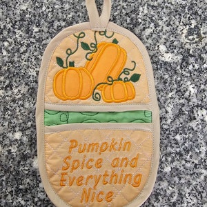 Könnte beinhalten: Beigefarbener, gesteppter Topflappen mit einem gestickten Design aus Kürbissen und Ranken in Grün und Orange. Der Text "Pumpkin Spice and Everything Nice" ist in Orange auf dem Topflappen gestickt.