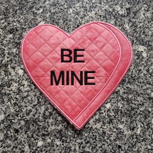 May include: A pink heart-shaped quilted hot pad with the text "BE MINE" embroidered in black.