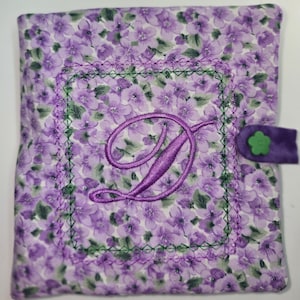 Puede incluir: Un artículo de tela cuadrado con un estampado floral morado. Cuenta con un borde verde cosido y una gran letra "D" morada bordada en el centro. Una solapa morada con un botón de flor verde asegura el artículo.