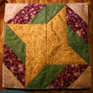 Puede incluir: Un bloque de colcha de retazos con un diseño de estrella en el centro. La estrella está hecha de una tela amarilla con un patrón floral. Las piezas circundantes están hechas de telas verdes, marrones y rosas con patrones florales.