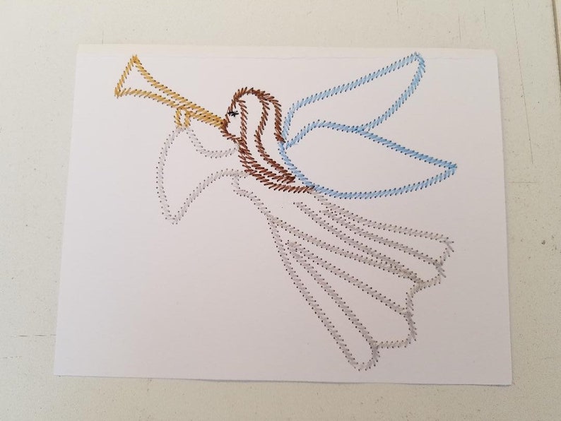 May include: A white card with a brown, blue, and yellow angel stitched on it. The angel is playing a trumpet.