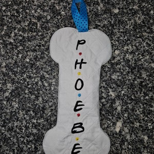 May include: A white, bone-shaped fabric item with the name "PHOEBE" embroidered in black, with colorful dots between the letters. It has a quilted pattern and a blue ribbon loop for hanging. The item is likely a decorative piece.