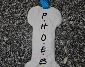 以下が含まれることがあります： 白い骨型の布製アイテムで、黒で「PHOEBE」という名前が刺繍されており、文字の間にカラフルなドットがあります。キルティング模様と、吊り下げ用の青いリボンループが付いています。装飾品である可能性が高いです。