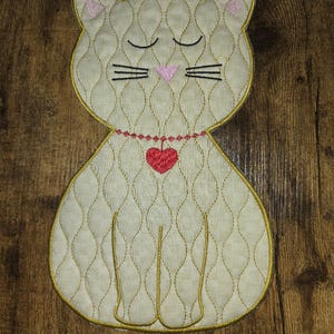 Pode incluir: Um gato branco acolchoado com os olhos fechados, um nariz rosa e um coração vermelho em uma corrente de ouro. O gato está sentado em uma superfície de madeira marrom.