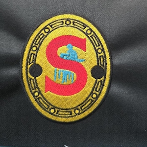 Può includere: Una toppa ricamata dorata con una lettera "S" rossa all'interno di un cerchio. La lettera "S" ha una figura blu seduta sopra.