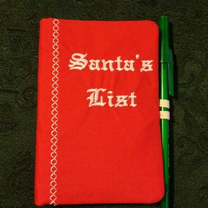 Peut inclure: Un carnet en tissu rouge avec des coutures blanches et les mots "Liste du Père Noël" brodés sur le devant. Un stylo vert est à côté du carnet.