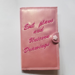 Peut inclure: Un carnet en satin rose avec une fermeture bouton en forme de fleur rose. Le carnet porte l'inscription "Evil plans and unicorn drawings" brodée sur le devant.