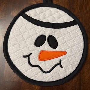 May include: White quilted pot holder with a black border and a snowman face embroidered on it. The snowman has black eyes, a black mouth, and an orange carrot nose.