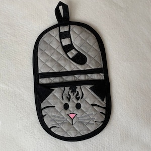 Pode incluir: Uma luva de forno acolchoada cinza com um design de gato preto. A cauda do gato está pendurada e seu rosto é visível com olhos pretos e um nariz rosa.