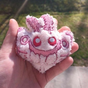 Könnte beinhalten: Ein rosa-weißes Plüschtier in Form eines Schmetterlings mit gestickten Details. Das Spielzeug hat große, weiße Augen mit roten Pupillen und ein rosafarbenes Lächeln.