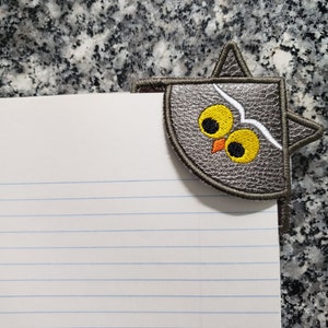 Op de afbeelding: Een grijze uilvormige bladwijzer met gele ogen en een witte snavel. De bladwijzer is gemaakt van vilt en is bevestigd aan een pagina van een genummerd notitieboek.