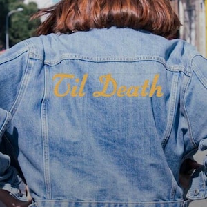 Puede incluir: Chaqueta vaquera azul claro con la frase bordada "Til Death" en amarillo en la espalda. La chaqueta tiene un diseño clásico con botones y bolsillos. El cabello de la persona es castaño rojizo.