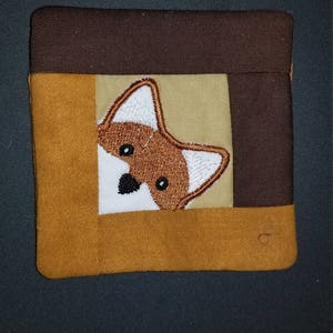 Puede incluir: Un cuadrado de tela marrón, beige y blanco con un zorro bordado que asoma por la esquina superior derecha.