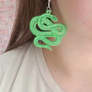 Puede incluir: Un par de pendientes de serpiente verde. Los pendientes están hechos de un material texturizado y tienen forma de serpiente enroscada sobre sí misma. Los pendientes cuelgan de ganchos plateados.