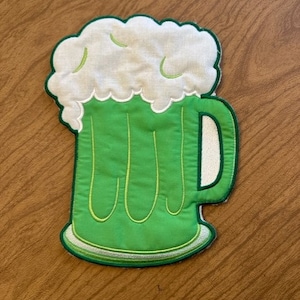 May include: A green and white felt beer mug with a foam top. The mug is embroidered with a green outline and white stitching.