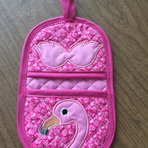 Könnte beinhalten: Rosa gesteppter Topflappen mit einem Flamingo-Motiv. Der Flamingo ist rosa mit einem schwarzen Auge und einem gelben Schnabel. Der Topflappen hat eine Schlaufe zum Aufhängen.