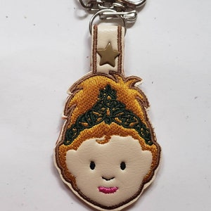 May include: A beige leather keychain with a gold star and an embroidered cartoon face with a green crown.