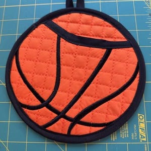 May include: Round orange and black quilted pot holder designed to resemble a basketball. The pot holder has a black loop for hanging and black stitching to create the basketball design. The pot holder is approximately 8 inches in diameter.