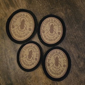 May include: Four circular patches with black borders and tan centers. The patches feature the text "THE SINGER AXE" around a central design of a brown axe. The patches are arranged on a wooden surface.
