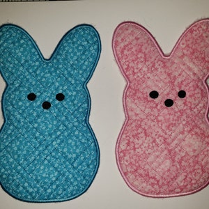Peut inclure: Deux maniques en forme de lapin de Pâques matelassées. L'une est bleue avec un imprimé floral blanc et l'autre est rose avec un imprimé floral blanc.
