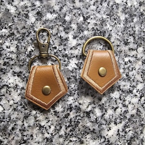 May include: Two brown leather keychains with a gold metal ring and a gold metal snap hook. The keychains are shaped like a hexagon with a button in the center.