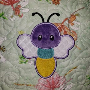 May include: A quilted square featuring a plush, embroidered butterfly. The butterfly has a purple head, white wings, a teal midsection, and a yellow body. The background is a light green fabric with floral and insect patterns.