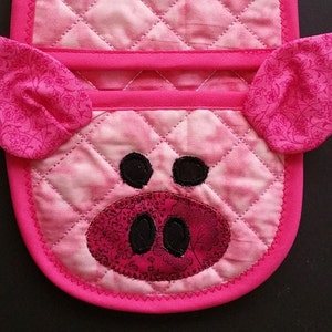 May include: Pink quilted hot pad with a pig face design. The pig has black eyes, a pink snout, and pink ears with a floral pattern.