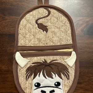 Op de afbeelding: Een bruine en witte ovenwant met een koe-ontwerp. De koe heeft een bruine neus, bruine ogen en bruin haar. De ovenwant heeft een lus om op te hangen.