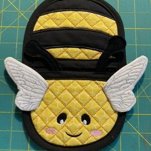 May include: A yellow and black bee pot holder with white wings and a smiling face. The pot holder is quilted with a honeycomb pattern.