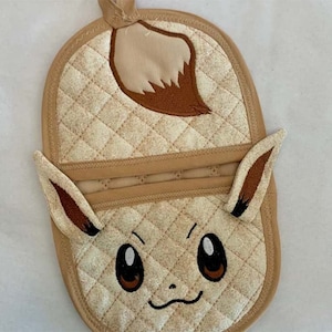Puede incluir: Una manopla de horno acolchada marrón y beige con un personaje de Eeveelution bordado. El personaje tiene orejas marrones, una cola marrón y una cara marrón con ojos blancos.