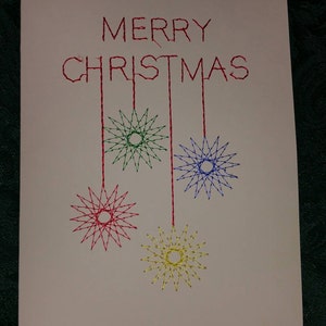 May include: A white card with the words "Merry Christmas" embroidered in red thread. Four embroidered stars hang from the words, one green, one blue, one red, and one yellow.