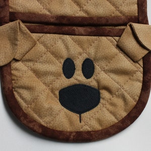 May include: Brown quilted oven mitt with a brown trim and a black embroidered dog face. The dog has two black eyes and a black nose.
