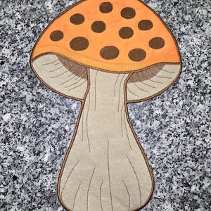 May include: Embroidered mushroom patch featuring an orange cap with brown spots and a tan stem. The patch has a brown outline and is likely for decorative purposes, such as on clothing or bags.