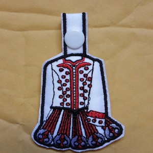 May include: White fabric patch with an embroidered image of a red and white Irish dance dress. The dress has a white bodice with red polka dots and a red skirt with black and white trim.