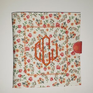 May include: A white floral patterned notebook cover with a orange embroidered monogram "CEJ" in a square frame.