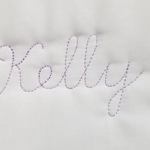 May include: A white fabric with the name "Kelly" embroidered in a cursive font with purple thread.