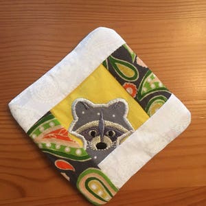 May include: A yellow, white and green fabric coaster with a grey embroidered raccoon in the centre.