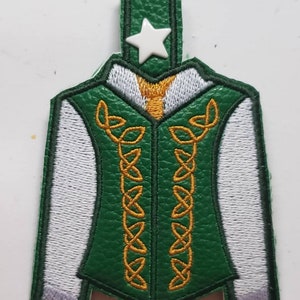 May include: A green and white embroidered jacket-shaped keychain with gold Celtic knot designs. The jacket has a white star on top and a gold collar. The sleeves are white, and the jacket is green.