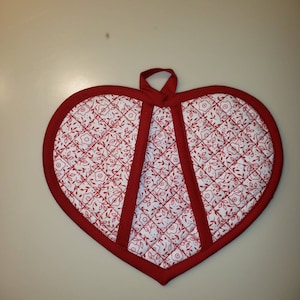 May include: Red heart-shaped pot holder with a white background and a floral pattern. The pot holder has a quilted design and a red trim.