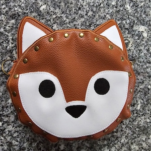 May include: A brown faux leather purse shaped like a fox's face with white accents and black eyes. The purse has gold-colored rivets around the edges.