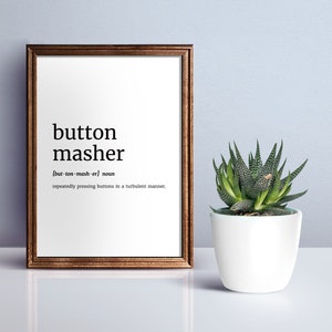 May include: A framed print with the definition of "button masher" in black text on a white background. The frame is brown. A small potted succulent plant sits to the right of the frame.